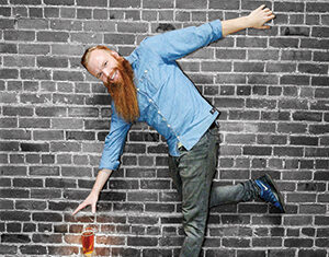 A man with a long red beard and blue shirt appears to be standing sideways on a brick wall, reaching down to touch a glass of beer placed against the wall. The image creates an illusion of defying gravity.
