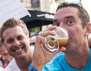 Two men smiling at an outdoor event; one in a blue shirt sips from a glass of beer while the other, in white, smiles at the camera. People and signs are blurred in the background.