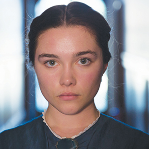 A young woman with fair skin and dark hair pulled back gazes directly at the camera with a serious expression, wearing a high-collared blue dress and standing in front of a blurred background with arched windows.