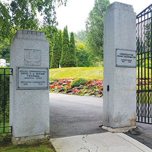 Open stone gates of a cemetery with plaques on each pillar, leading to a path surrounded by green grass, trees, and colorful flower beds in the background.