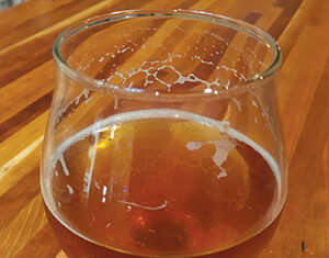 A stemmed glass filled with amber-colored beer sits on a wooden table, with foam residue visible on the inside of the glass.