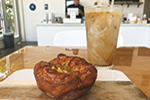 A close-up of a pastry on a plate and an iced coffee on a wooden table in a café, with a person at the counter in the blurred background.