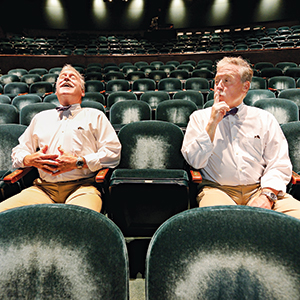 Two men wearing light shirts and bow ties sit in an empty theater. One man laughs with his head back, while the other holds a finger to his lips, gesturing for silence. Rows of empty green seats surround them.