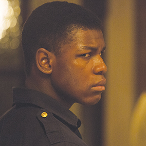 A young man with short hair, wearing a dark shirt with a gold button, looks over his shoulder with a serious and intense expression. The background is softly lit and slightly out of focus.