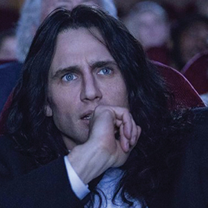 A man with long dark hair and blue eyes sits in a theater seat, looking intently ahead with his hand resting on his chin. The background is out of focus, showing other audience members.