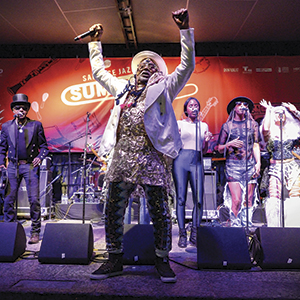 A lively band performs on stage at a festival, with a lead singer in a white hat and jacket raising an arm, surrounded by musicians and singers under colorful stage lights and a bright red banner.