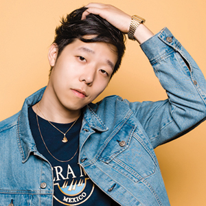 A young man wearing a denim jacket and black shirt poses with one hand running through his hair against a plain peach background. He wears a gold watch and necklace, and looks calmly at the camera.