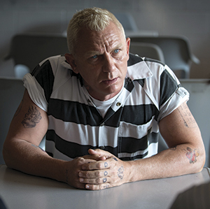 A man with short blond hair and tattoos on his hands sits at a table wearing a black-and-white striped prison uniform, looking serious in a room with gray chairs.