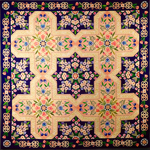 Intricate quilt with a symmetrical floral pattern in pink, green, and blue on cream and dark blue backgrounds, featuring cross-shaped designs and a decorative border with interlocking loops and small flowers.