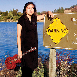 A woman in a black dress stands by a lake, holding a ukulele and leaning on a sign that says WARNING. She looks serious, with trees and blue water in the background.