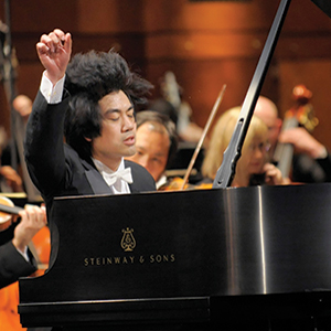 A pianist in a tuxedo passionately plays a grand Steinway & Sons piano on stage, with an orchestra performing in the background. His eyes are closed and one arm is raised, showing intense emotion.