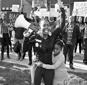 A Black woman holding a megaphone raises her fist while a young girl hugs her waist. They stand among a crowd holding Black Lives Matter signs at a protest.