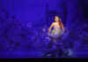A performer dressed as a mermaid sings on stage, surrounded by purple lighting and floating bubble effects, with a castle backdrop in the distance.