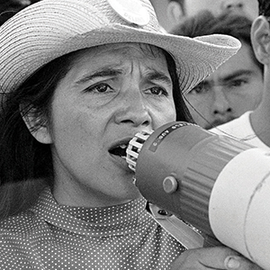 A woman wearing a hat uses a megaphone to speak, surrounded by people. The image is black and white and the woman appears to be passionately addressing a crowd.