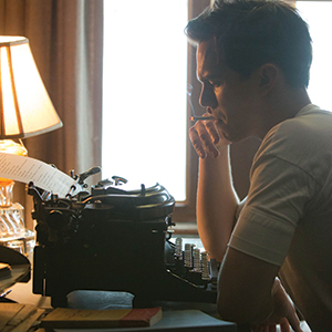A person in a white shirt sits at a desk by a window, holding a cigarette and looking thoughtfully at a typewriter with a sheet of paper. A lit lamp and books are also visible on the desk.