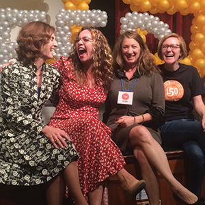 Four women sit closely together, smiling and laughing in front of a backdrop made of silver and gold balloons. They appear to be enjoying a celebratory event. One wears a badge, another has a shirt with 150 on it.