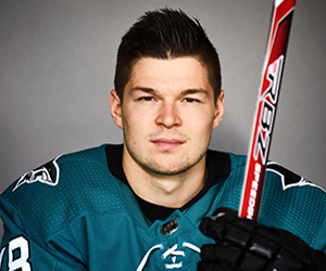 A male hockey player in a teal jersey holds a red and white hockey stick upright, looking directly at the camera against a neutral background.