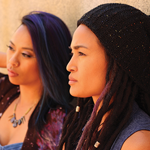 Two women with long hair, one wearing a black knit hat and dreadlocks, both looking thoughtfully into the distance against a sunlit beige wall.