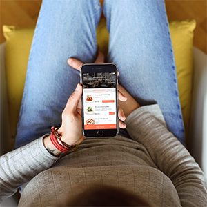 A person in jeans and a gray sweater sits on a chair, holding a smartphone displaying a food delivery app with meal options, viewed from above.