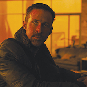 A man with short hair and a bruise on his face, wearing a dark jacket with a shearling collar, sits indoors in warm yellow-orange lighting, looking thoughtfully to the side.