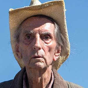 An elderly man wearing a straw hat and brown jacket looks ahead with a serious expression against a clear blue sky.