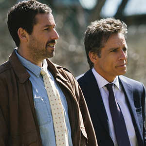 Two men stand side by side outdoors. One wears a brown jacket, light blue shirt, and polka dot tie; the other wears a dark suit and tie. Both look off into the distance with serious expressions.