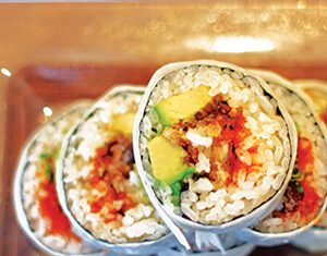 Four sushi burritos, wrapped in seaweed and foil, are sliced in half to reveal rice, avocado, vegetables, and other colorful fillings, arranged on a clear rectangular plate.