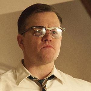 A man with short brown hair and glasses, wearing a white shirt and a striped tie, looks serious and determined in a softly lit room.