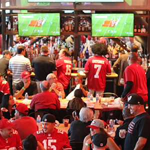 A lively sports bar filled with people wearing red football jerseys, watching a game on two large screens above the bar, eating, drinking, and socializing.