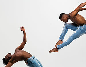 Two shirtless men wearing jeans perform dynamic dance moves; one leaps high in the air with knees bent, while the other leans backward, balancing on one leg, against a plain white background.