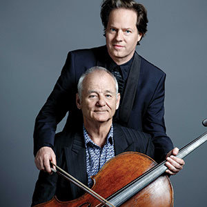Two men pose for a portrait against a gray background. The seated man holds a cello and bow, while the standing man rests his hands on the seated mans shoulders. Both wear dark jackets and look toward the camera.