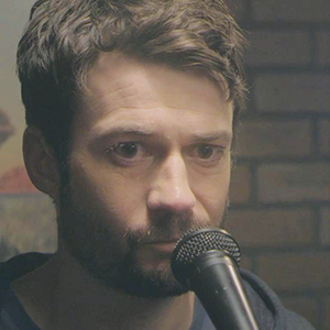 A man with short brown hair and a beard stands close to a microphone, looking serious. There is a brick wall and part of a painting in the blurred background.