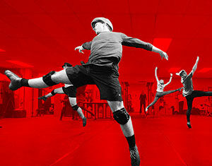 Four dancers leap energetically in a dance studio. The background is tinted red, highlighting their dynamic poses. The dancers wear casual athletic clothing and knee pads, emphasizing movement and action.
