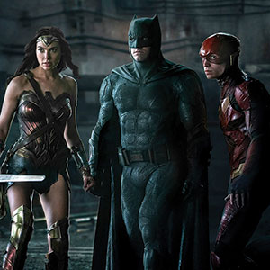 Three superheroes—Wonder Woman, Batman, and The Flash—stand together in a dark, industrial setting, looking alert and ready for action in their distinctive costumes.
