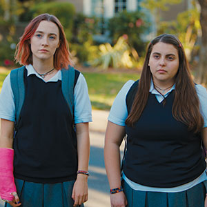 Two teenage girls in school uniforms stand side by side outdoors. One has shoulder-length pink hair and a pink cast on her arm; the other has long brown hair. Both have neutral expressions. Trees and a house are in the background.