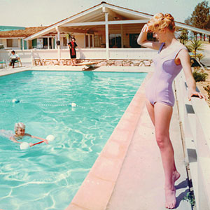 A woman in a lavender swimsuit stands by the edge of an outdoor swimming pool, shielding her eyes from the sun. Another person swims with a float, and a third person is seen in the background near a mid-century motel.