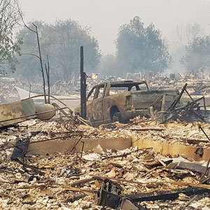 A burned-out truck sits amid the charred remains of a destroyed neighborhood, surrounded by debris and ash, with smoke and fire still visible in the background.