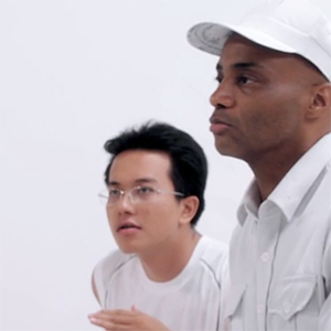 Two people in white outfits stand against a plain white background. One wears glasses and looks forward attentively; the other wears a white cap, shirt, and looks ahead with a serious expression.