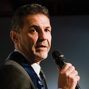 A man in a suit and tie speaks into a microphone. He has short, dark hair with some gray and stands in front of a blurred, dark background.