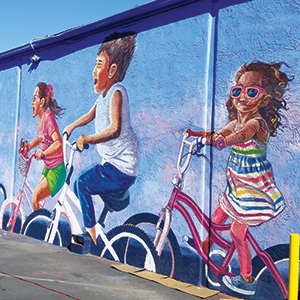 A colorful mural on a wall shows three children riding bicycles, with one girl wearing sunglasses and a striped dress. The background is blue, giving a lively and playful atmosphere.