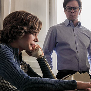 A woman with short hair speaks on a corded telephone, looking concerned, while a man in glasses and a button-up shirt stands behind her, watching intently in a softly lit room.
