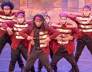 A group of six dancers in matching red and gold jackets and black pants perform a hip-hop routine on stage, striking dynamic poses with serious expressions.