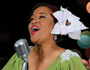 A woman in a green dress sings passionately into a vintage microphone. She wears a large white flower in her hair and has one arm raised, performing against a dark, blurred background.