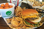 A cheeseburger with lettuce, tomato, and onions sits on a tray with crispy onion rings. Beside it is a cup of tater tots and a container of ketchup. The food appears to be from Wahlburgers.