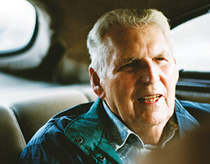 An elderly man with gray hair sits in the backseat of a car, wearing a teal jacket over a shirt, softly smiling and talking, with sunlight filtering in through the window.