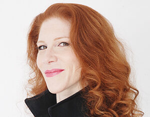 A woman with long, curly red hair and fair skin smiles slightly, wearing a sleeveless black top with a high collar against a plain white background.