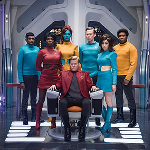 Seven people in colorful, retro sci-fi uniforms pose on a spaceship bridge. One man sits in a central captain’s chair, while six others stand around him against a futuristic backdrop with control panels and lights.