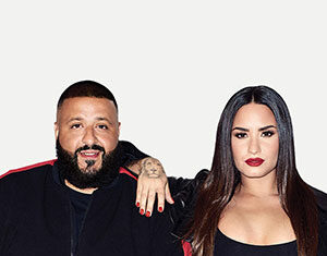 A man with a beard and a woman with long dark hair pose together, both wearing black outfits. The woman rests her arm on the mans shoulder against a plain white background.