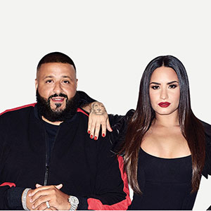 A man with a beard and a woman with long dark hair pose together, both wearing black outfits. The woman rests her arm on the mans shoulder against a plain white background.