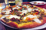 A close-up of a freshly baked pizza topped with melted cheese, ground meat, and colorful peppers on a white plate. A drink and another plate are visible in the blurred background.